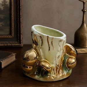 May include: A ceramic vase with a pale cream and green glaze, featuring a textured surface adorned with multiple three-dimensional mushroom shapes finished in a reflective metallic gold luster. The vase has an asymmetrical, slanted top opening.