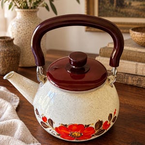May include: A cream-colored enamel tea kettle with a speckled finish, featuring a dark brown lid and handle. The body is decorated with a vibrant red floral pattern with brown leaves. The kettle has a curved spout and a matching brown knob on top.