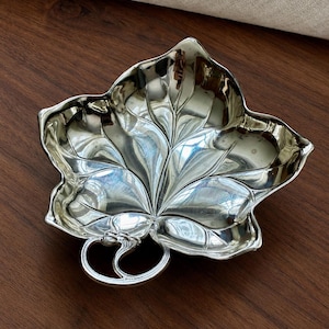 May include: A polished silver-toned metal dish shaped like a large maple leaf. The dish features embossed vein details across the surface and a curved, loop-like handle at the base, creating a reflective, smooth, and metallic appearance.