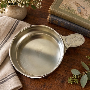 May include: A round, silver-toned metal dish with a smooth, reflective interior surface and a single handle extending from the side. The handle features an embossed, detailed illustration of a rabbit in a jacket, captured in a running pose.