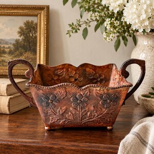 May include: A rectangular metal bowl with a copper-toned finish, featuring embossed floral patterns on the sides, a scalloped top edge, and two curved handles on the ends. The bowl stands on four small feet and has a textured, weathered appearance.