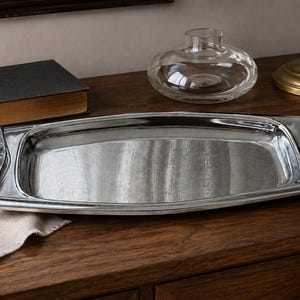 May include: A rectangular silver-toned metal serving tray with a polished, reflective surface and curved edges. The tray features integrated handles on both ends, each designed with a series of three horizontal cutouts for a decorative finish.