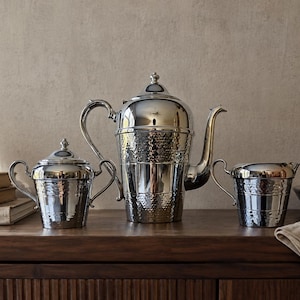 May include: A three-piece silver-toned metal tea set consisting of a large lidded teapot, a smaller lidded sugar bowl, and an open creamer pitcher. Each piece features a polished finish with a distinct hammered texture on the lower body.