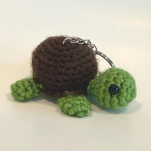 May include: A handmade crochet turtle keychain. The turtle has a brown body, green head and limbs, and a black eye. The keychain is attached with a silver metal ring and chain.