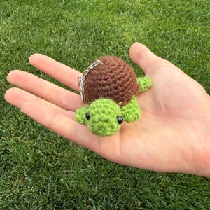 May include: A handmade crochet turtle keychain. The turtle has a brown shell and green head, feet, and tail. The keychain is small and compact, perfect for attaching to keys or a backpack.