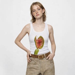 May include: White vest top with a colourful graphic of a Venus flytrap. The plant is shown with red and green hues, with water droplets. The vest top is worn with a brown belt and khaki trousers.