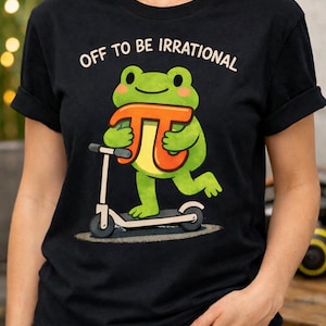 May include: Black t-shirt featuring the text "OFF TO BE IRRATIONAL." A green frog with a Pi symbol rides a scooter. The Pi symbol is orange and yellow. The frog has pink cheeks. The t-shirt has rolled-up sleeves.