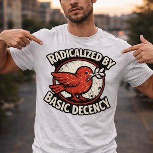 Puede incluir: Camiseta gris claro con un gráfico de un pájaro rojo sosteniendo una rama de olivo, rodeado por el texto "RADICALIZED BY BASIC DECENCY". La camiseta es de material suave.