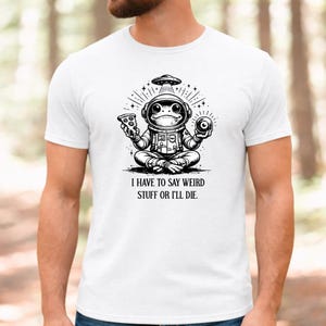 Puede incluir: Una camiseta blanca con un gráfico negro de una rana con traje de astronauta sentada en posición de loto. La rana sostiene una porción de pizza y un objeto redondo, con un OVNI encima. El texto debajo dice: I HAVE TO SAY WEIRD STUFF OR I'LL DIE.
