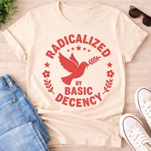 Puede incluir: Camiseta color crema con el texto rojo "RADICALIZED BY BASIC DECENCY" que rodea una paloma roja con una rama de olivo. El diseño incluye elementos florales y de estrellas. La camiseta se muestra con vaqueros azules y zapatillas blancas.