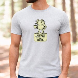 Puede incluir: Una camiseta gris jaspeado claro con un gráfico de un sapo verde sosteniendo una taza de café. Debajo del sapo, un recuadro rectangular amarillo contiene el texto en negro: I HAVE TO SAY WEIRD STUFF OR I'LL DIE.