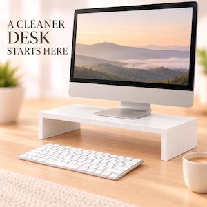 Op de afbeelding: Een witte rechthoekige monitorstandaard met een gladde afwerking, geplaatst op een houten oppervlak. Een computermonitor staat op de standaard en toont een berglandschap. De tekst A CLEANER DESK STARTS HERE is linksboven zichtbaar.