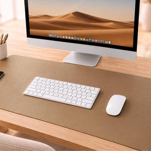 May include: A large, rectangular, tan-coloured desk mat with a smooth, matte surface. The mat is positioned on a wooden desk surface, serving as a base for a white wireless keyboard and a white wireless computer mouse, creating a clean workspace.