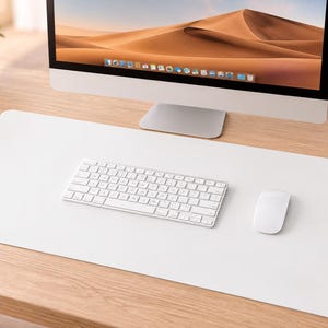 Puede incluir: Una alfombrilla de escritorio grande, rectangular y de color blanco sólido con esquinas redondeadas, colocada sobre una superficie de madera clara. Un teclado inalámbrico blanco y un ratón inalámbrico están sobre la alfombrilla, frente al monitor.