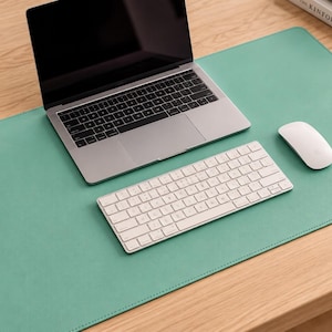 Puede incluir: Una alfombrilla de escritorio grande y rectangular de color verde menta con bordes cosidos. La alfombrilla proporciona una superficie lisa para un portátil plateado, un teclado inalámbrico blanco y un ratón inalámbrico blanco sobre una mesa.