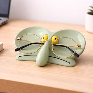 May include: A light green ceramic glasses holder shaped like a cartoon character's face, with yellow eyes and a long nose. Gold-rimmed glasses rest on the holder. The holder sits on a wooden desk.