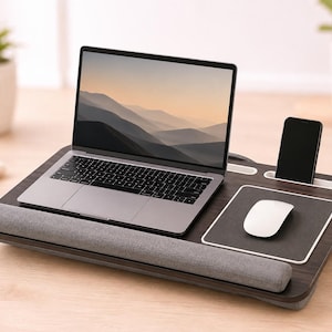 Puede incluir: Un escritorio para portátil de madera oscura con reposamuñecas de tela gris, alfombrilla de ratón negra integrada, ratón blanco, ranura para smartphone y portalápices. El escritorio sostiene un portátil plateado con un paisaje de montaña.
