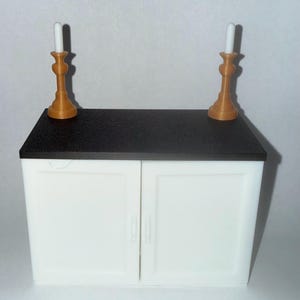 Peut inclure: Un petit meuble blanc avec un plan de travail noir. Deux bougeoirs dorés avec des bougies blanches sont posés dessus. Le meuble a deux portes avec des poignées argentées. Le design est simple et élégant.