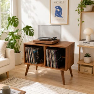 Könnte beinhalten: Ein Plattenspieler-Ständer im Mid-Century-Modern-Stil aus Holz. Der Ständer hat zwei offene Fächer, die mit Schallplatten gefüllt sind. Auf der Oberseite befindet sich ein Plattenspieler mit einer transparenten Abdeckung. Der Raum ist mit Pflanzen und einem gerahmten Kunstdruck dekoriert.