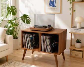 May include: A mid-century modern record player stand made of wood. The stand has two open compartments filled with vinyl records. A turntable with a clear dust cover sits on top. The room is decorated with plants and a framed art print.