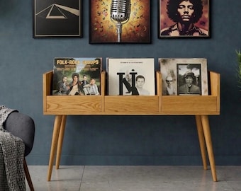 May include: A wooden record storage unit with three compartments, holding vinyl records. The unit has angled legs and is positioned beneath framed art. The art includes a microphone, a portrait, and a geometric design.