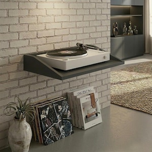 May include: A white turntable with a black record sits on a gray floating shelf against a brick wall. Below, a stack of vinyl records and books with a leather bookmark are visible. A decorative vase with a plant is also present.