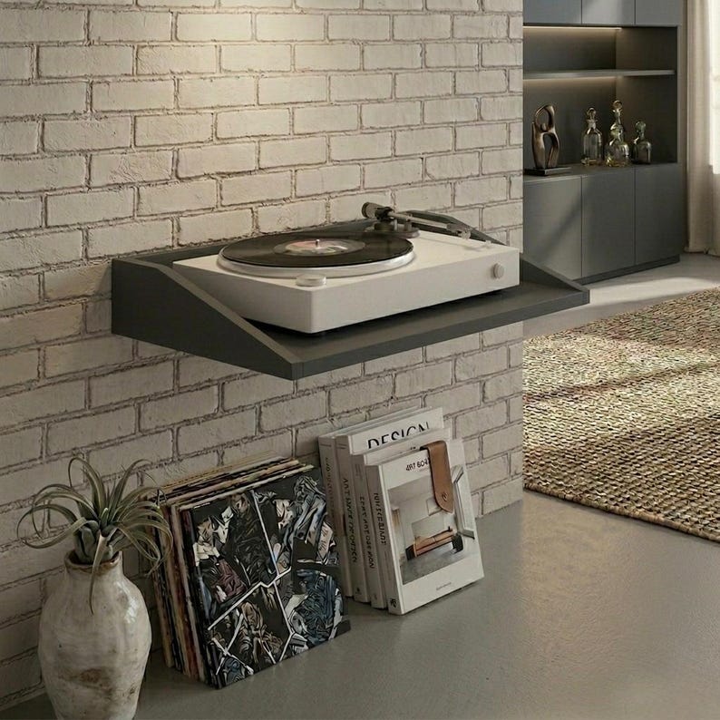 May include: A white turntable with a black record sits on a gray floating shelf against a brick wall. Below, a stack of vinyl records and books with a leather bookmark are visible. A decorative vase with a plant is also present.