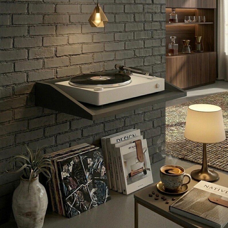 May include: A white turntable with a black vinyl record sits on a dark gray shelf mounted on a brick wall. A stack of vinyl records and books are below the shelf. A small lamp and coffee cup are on a side table.