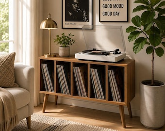 May include: A mid-century modern record player stand with a white turntable and a collection of vinyl records. The wooden stand has multiple compartments for record storage. A gold-toned lamp and framed art pieces are visible in the background, creating a cozy living room setting.