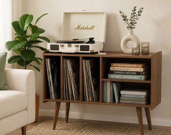 May include: A mid-century modern record player stand with a white Marshall record player on top. The stand is made of brown wood and has compartments for vinyl records and books. A green plant and a white vase with greenery are also visible.