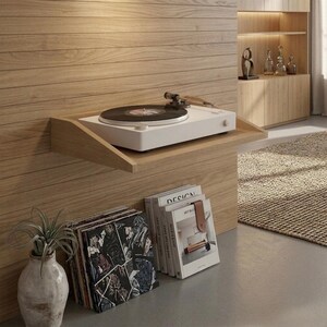 May include: A white turntable with a black vinyl record sits on a wooden shelf. The shelf is mounted on a light wood-paneled wall. Below, a stack of vinyl records and books are visible, along with a decorative vase with a plant.