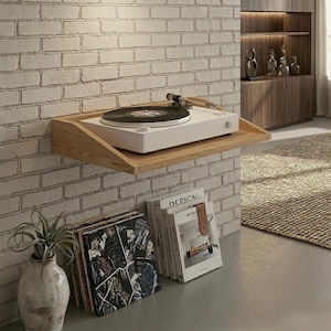 May include: A white turntable with a black record sits on a wooden shelf mounted on a brick wall. Below, a stack of vinyl records and books are visible. A decorative vase with a plant is on the left.