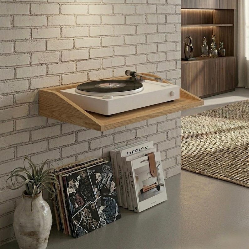 May include: A white turntable with a black record sits on a wooden shelf mounted on a brick wall. Below, a stack of vinyl records and books are visible. A decorative vase with a plant is on the left.