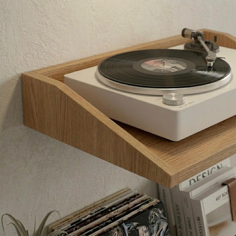 May include: A white turntable with a black vinyl record sits on a light wood shelf. The shelf is mounted on a white wall. Below the shelf are stacks of vinyl records and books. The turntable has a silver tone arm and control knob.