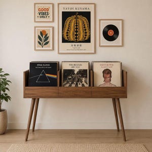 Può includere: Un mobile in legno per riporre dischi con gambe angolate contiene tre album in vinile. Sopra, opere d'arte incorniciate includono una stampa di zucca Yayoi Kusama, un disco e altre due stampe. Il testo "GOOD VIBES ONLY" è visibile.