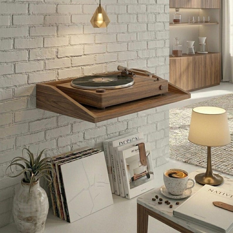 May include: A wooden record player mounted on a wall shelf. The turntable is brown with a black record on it. The shelf is mounted on a white brick wall. A small lamp and books are on a table below.