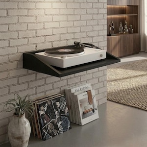 May include: A white turntable with a black record sits on a black shelf mounted on a white brick wall. Below, a stack of vinyl records and books with a plant in a vase add to the decor. The shelf is approximately 12 inches deep.