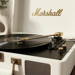 May include: A white Marshall record player with a black vinyl record playing. The record player has gold accents, including the brand name and knobs. The record player is open, and the needle is on the record.
