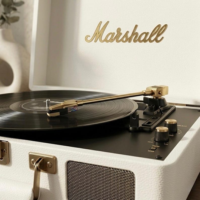 May include: A white Marshall record player with a black vinyl record playing. The record player has gold accents, including the brand name and knobs. The record player is open, and the needle is on the record.