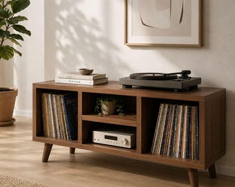 May include: A rectangular dark wood media console with four tapered legs, featuring two open side compartments filled with vinyl records, a central shelf holding a silver audio receiver, and a top surface supporting a black turntable and books.