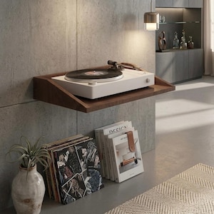 May include: A white turntable with a black record sits on a dark wood shelf mounted on a gray concrete wall. Below, a stack of vinyl records and books are arranged near a decorative vase with a plant. A modern, minimalist interior.