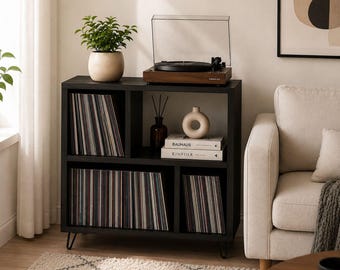 May include: A black wooden storage unit with hairpin legs featuring three open compartments for vinyl records, a top surface holding a brown turntable with a clear lid, a potted green plant, a ceramic vase, and two books labeled Bauhaus and Kinfolk.