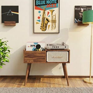 May include: A mid-century modern style record player stand with a built-in speaker. The stand is made of dark wood and has angled legs. A framed "Blue Note Jazz" poster hangs above the stand, with vinyl records on display.