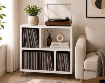 May include: A white wooden storage unit with four compartments, supported by black metal hairpin legs. The unit holds vinyl records, a brown wooden turntable, a green potted plant, a brown glass bottle with reeds, and two books labeled Bauhaus and Kinfolk.