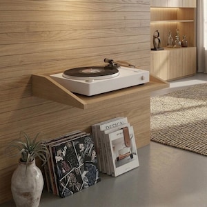 May include: A white turntable with a black vinyl record sits on a wooden shelf. The shelf is mounted on a light wood-paneled wall. Below, a stack of vinyl records and books are visible, along with a decorative vase with a plant.
