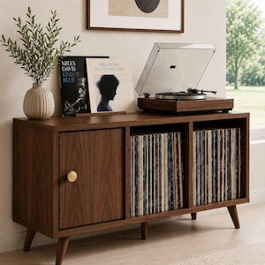Puede incluir: Una consola multimedia de madera oscura con patas cónicas, que cuenta con una puerta de armario con un pomo redondo dorado y dos compartimentos de almacenamiento abiertos llenos de discos de vinilo. Un tocadiscos descansa sobre la parte superior.