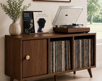 May include: A dark wood media console with tapered legs, featuring one cabinet door with a round gold knob and two open storage compartments filled with vinyl records. A record player sits on top, alongside a potted plant and two vinyl album covers.