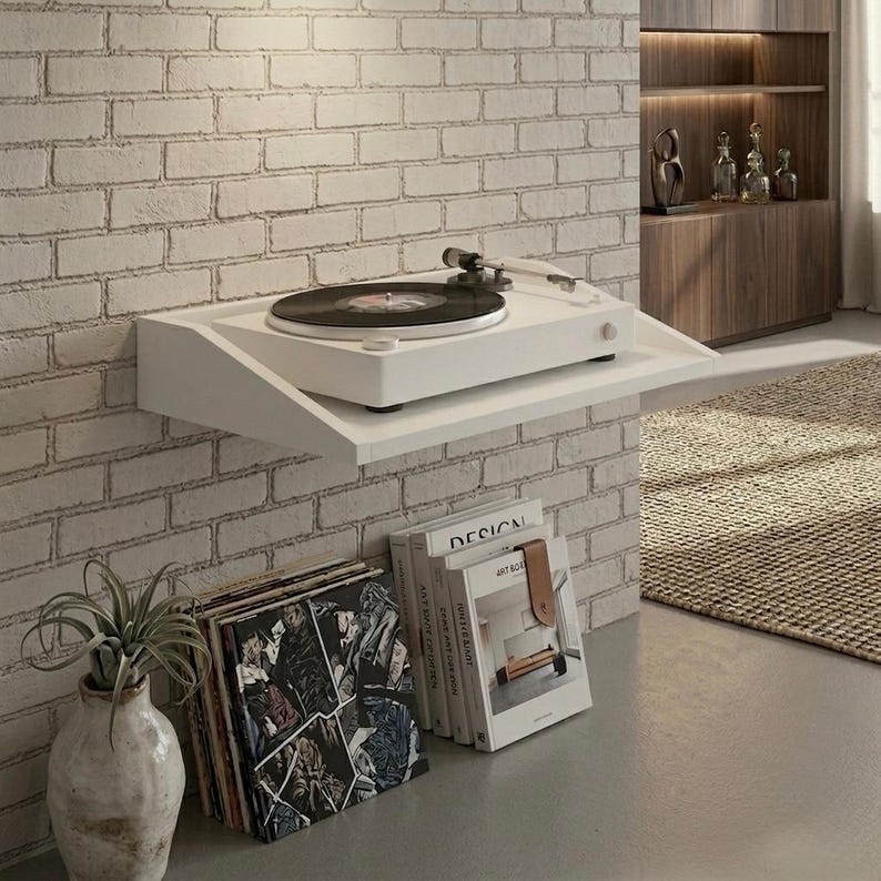 May include: A white turntable with a black vinyl record sits on a white shelf mounted on a brick wall. A stack of vinyl records and books are below the shelf. A decorative vase with a plant is on the left.
