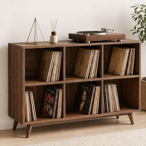 May include: A mid-century modern style record player and vinyl storage unit. The dark brown wooden cabinet has six cubby holes filled with vinyl records. A turntable sits on top, along with a gold geometric decoration and a diffuser.