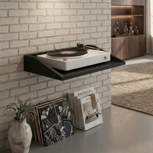 May include: A white turntable with a black record sits on a black shelf mounted on a white brick wall. Below, a stack of vinyl records and books with a plant in a vase add to the decor. The shelf is approximately 12 inches deep.